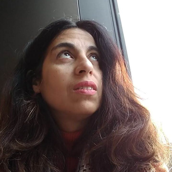 A woman, Dr Adriana Wilde, with long brown hair, is looking up beside a window on a sunny day. She is wearing an orange cardigan and a scarf in pastel colours.