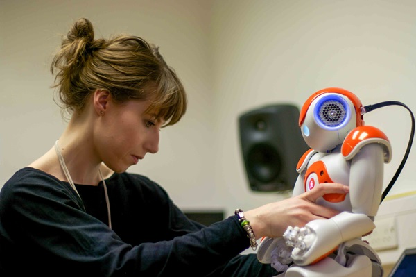 A woman with light brown hair tied back is interacting with a medium-sized orange and white humanoid robot in a classroom.