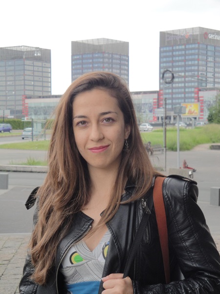 A smiling woman, Dr Dimitra Georgiadou, with long light brown hair, wearing an abstract-printed blouse in grey, blue, and yellow, along with a black leather jacket.